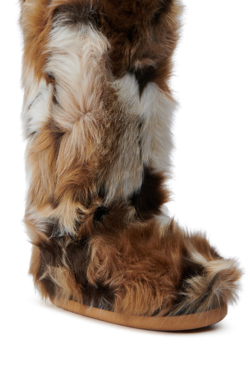 The image shows a close-up of the AZALEA WANG FENNEC MULTI FUZZY FLAT KNEE HIGH BOOT, crafted from soft, fluffy, multi-colored faux fur. The synthetic fur features patches of brown, white and black. The boot is designed with a rounded toe and a flat sole.