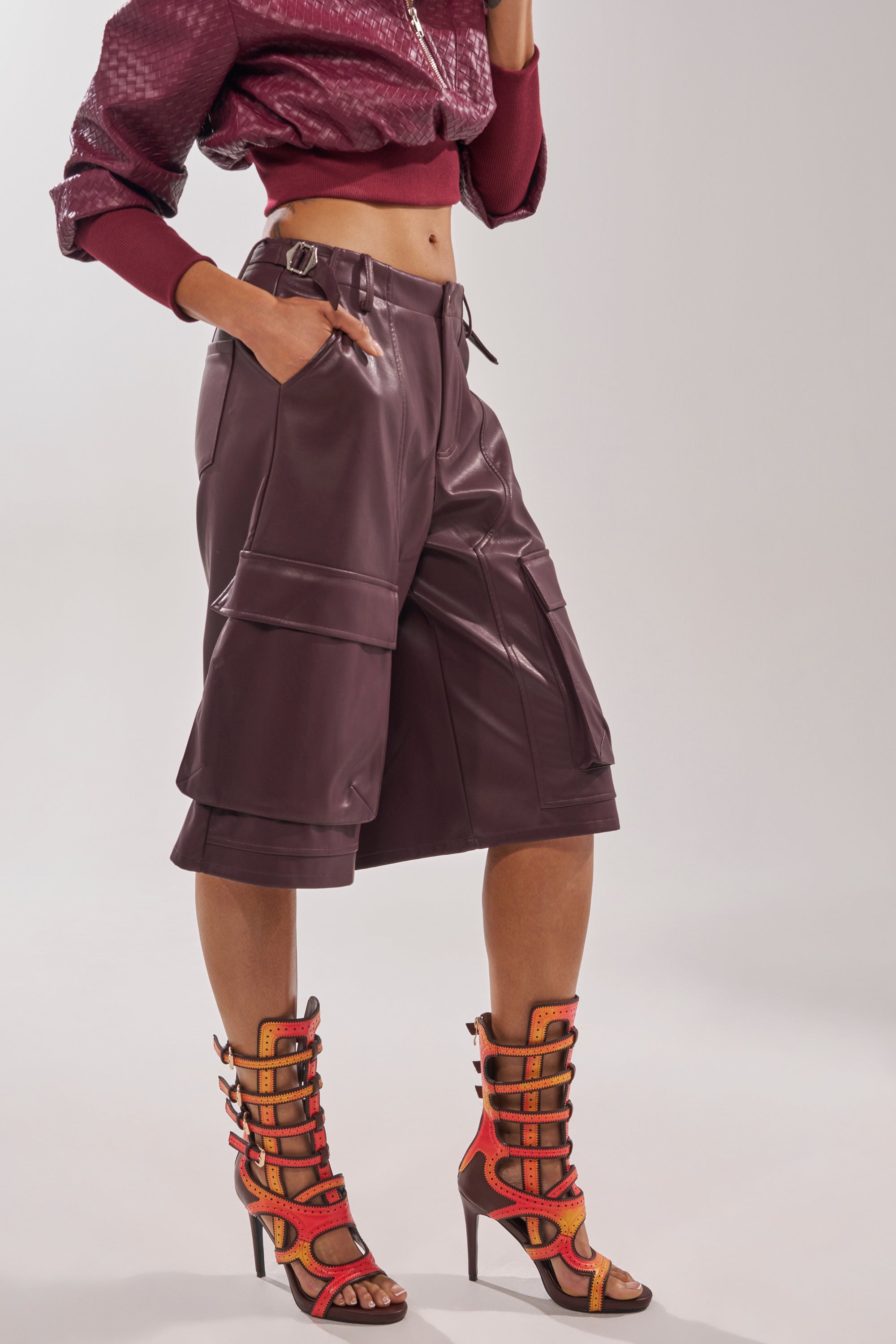 A person wearing a cropped maroon jacket, high-waisted maroon leather shorts, and AKIRA GOLD FEVER ORANGE MULTI OMBRE OPEN TOE BOOTIE poses with hands in pockets against a plain background. 1