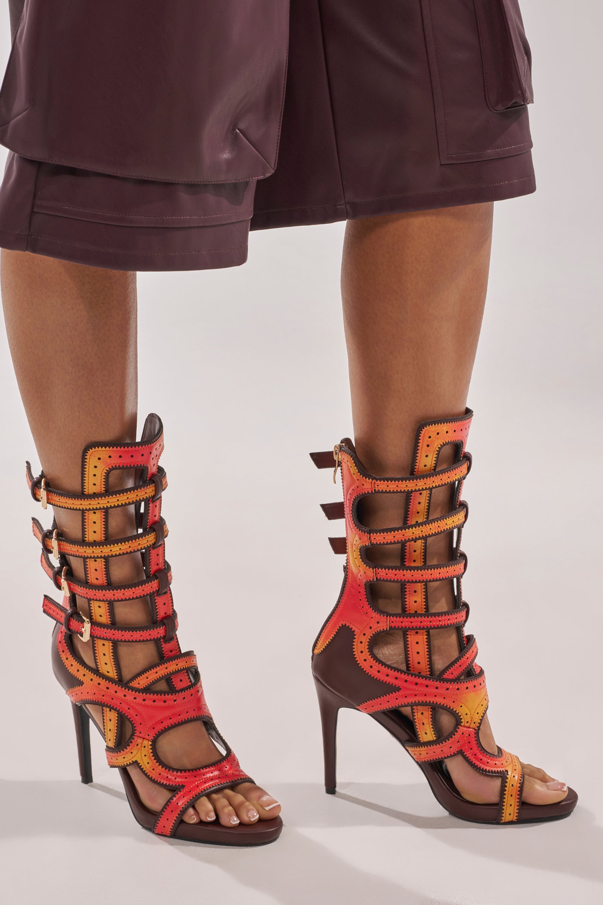 A person wearing burgundy shorts and the AKIRA GOLD FEVER ORANGE MULTI OMBRE OPEN TOE BOOTIE, featuring vibrant shades of red, orange, and yellow with multiple straps and cutout details. 2