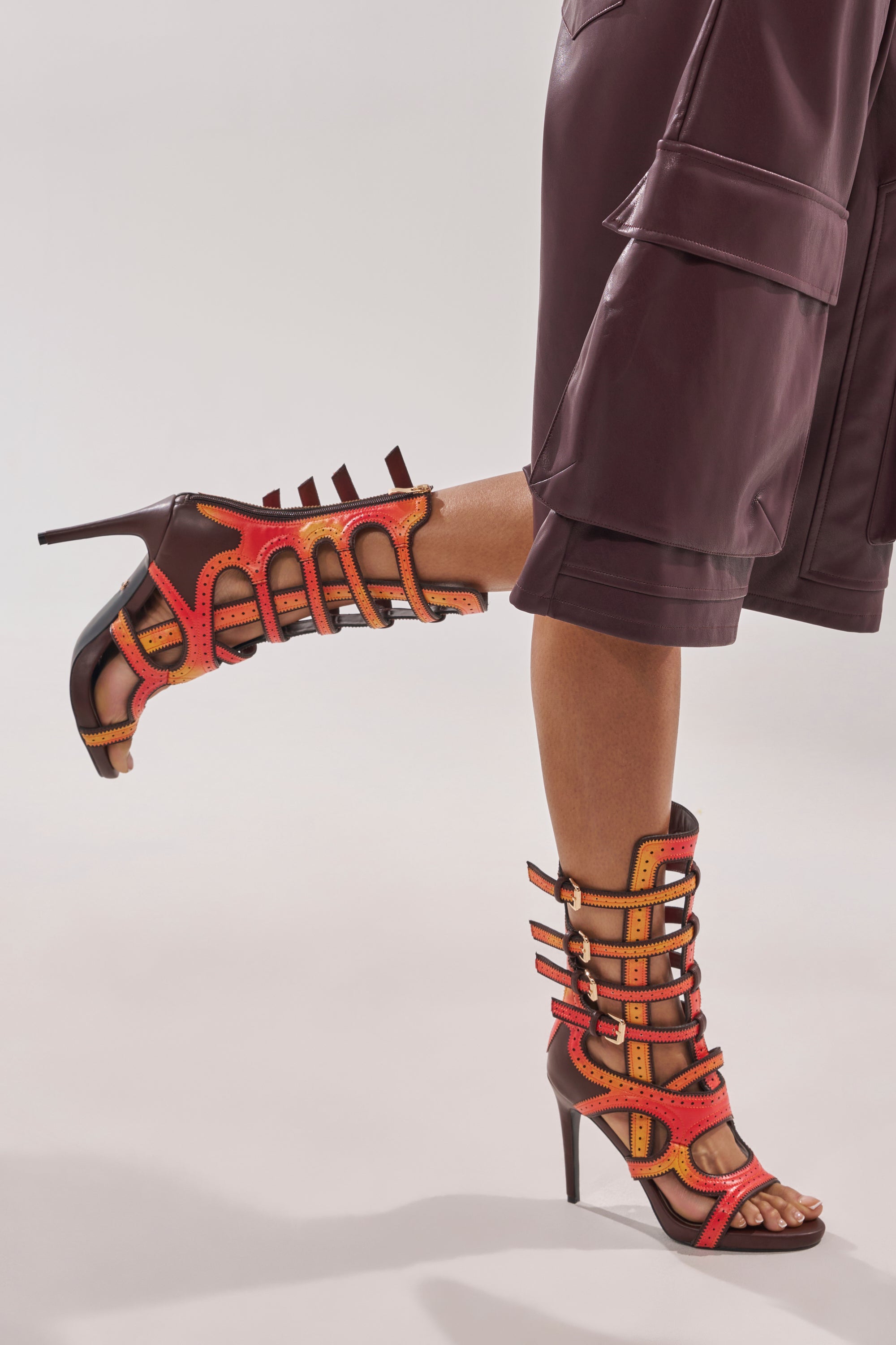 A person in burgundy knee-length shorts lifts one leg, highlighting the AKIRA GOLD FEVER ORANGE MULTI OMBRE OPEN TOE BOOTIE with vibrant red, orange, and brown straps and gold buckles against a neutral background. 0