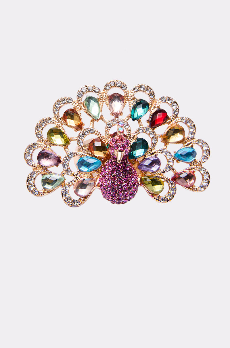 The PERRY COCKTAIL RING features a peacock design with a purple body and multicolored gem-studded tail feathers, set in gold tones and accented by clear stones, shown against a plain white background.