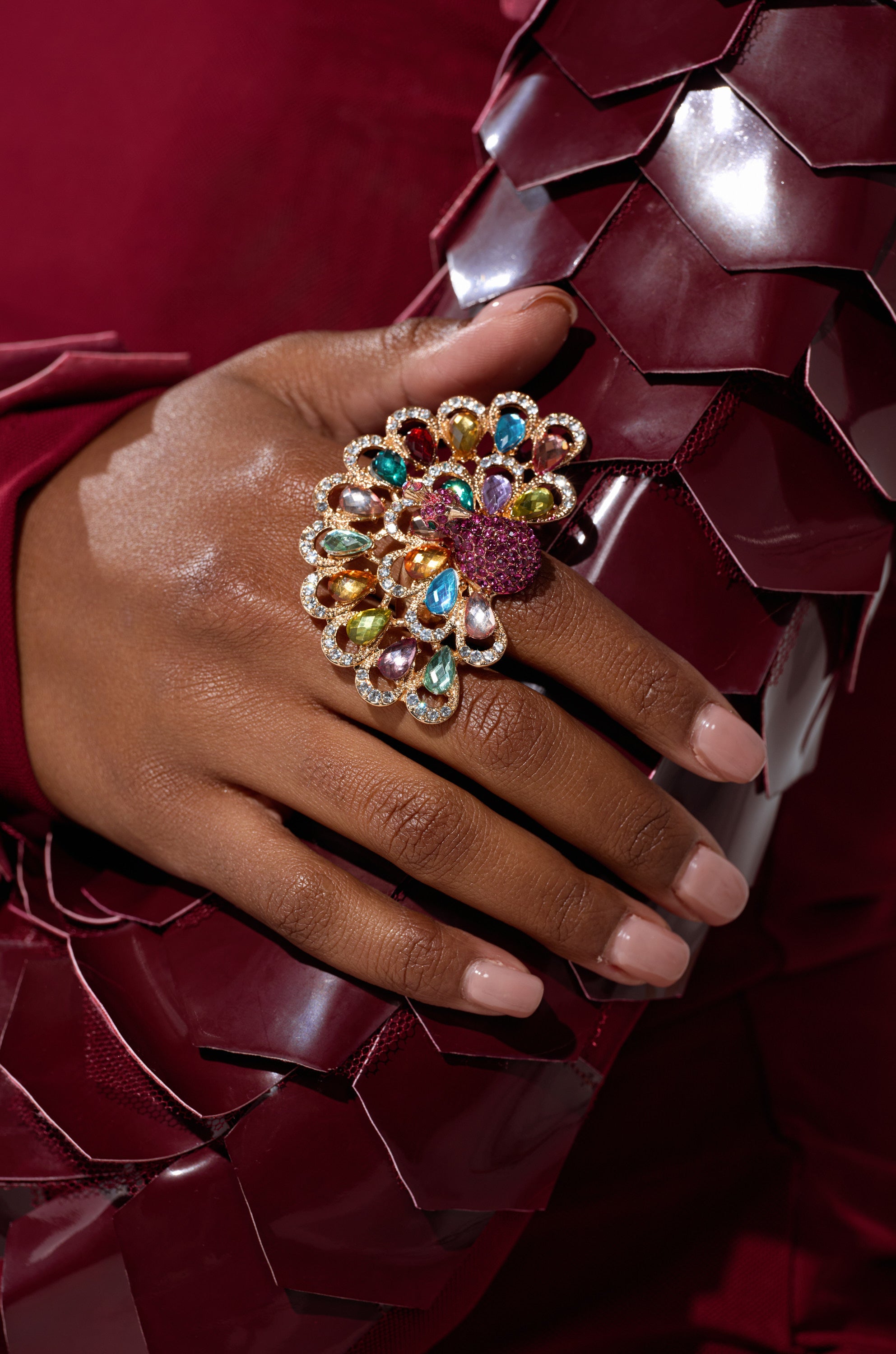 A hand with manicured nails wears the PERRY COCKTAIL RING, a large, colorful flower-shaped accessory with rhinestone accents, resting on shiny, dark red fabric with a scale-like texture. 1