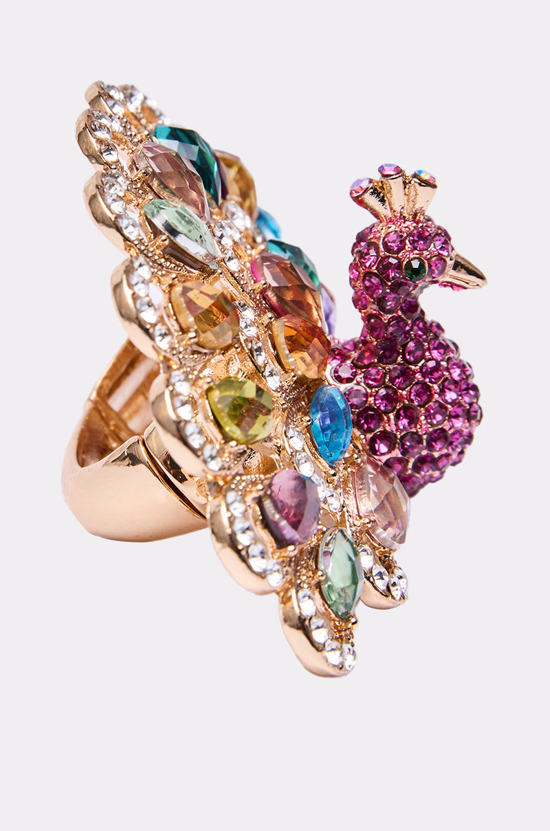 The PERRY COCKTAIL RING, shaped like a peacock and adorned with vibrant gemstones, pink stones on the body, and sparkling rhinestones, is showcased against a simple light background. 2
