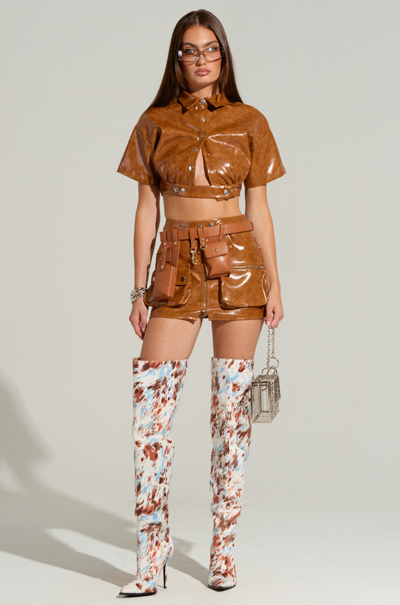 A woman stands against a neutral backdrop wearing a brown faux leather crop top and matching high-waisted mini skirt with attached pockets. She holds a small handbag and wears knee-high, white and brown patterned boots. With long hair, glasses, bracelets, and the stylish UTILITY BELT featuring an adjustable buckle, she exudes fashion-forward style.