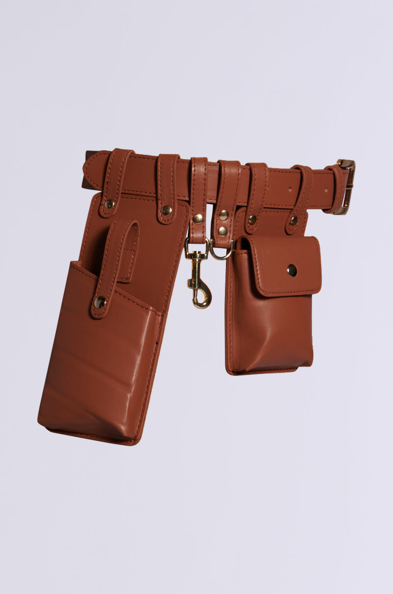 The UTILITY BELT, crafted from brown faux leather, features multiple compartments including two large pockets, a smaller pouch with a button closure, and a central metal clip. Its adjustable buckle ensures it fits comfortably while providing secure and organized storage.