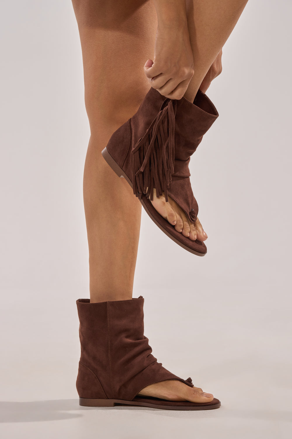 A person balances on one leg against a neutral background, bending down to adjust the AZALEA WANG FIDDA CHOCOLATE GLADIATOR FLAT SANDAL with open toes and brown faux suede fringe on their raised foot. 0