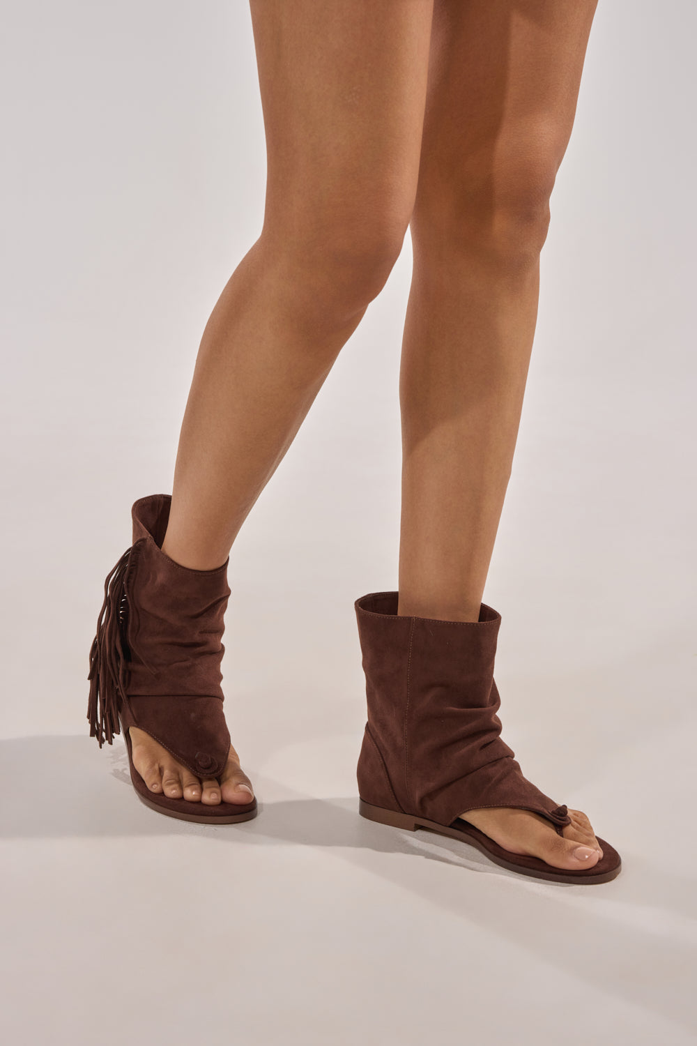 Only the legs and feet are visible as a person stands on a neutral background, wearing the AZALEA WANG FIDDA CHOCOLATE GLADIATOR FLAT SANDAL with slouchy, boho fringe details. 2