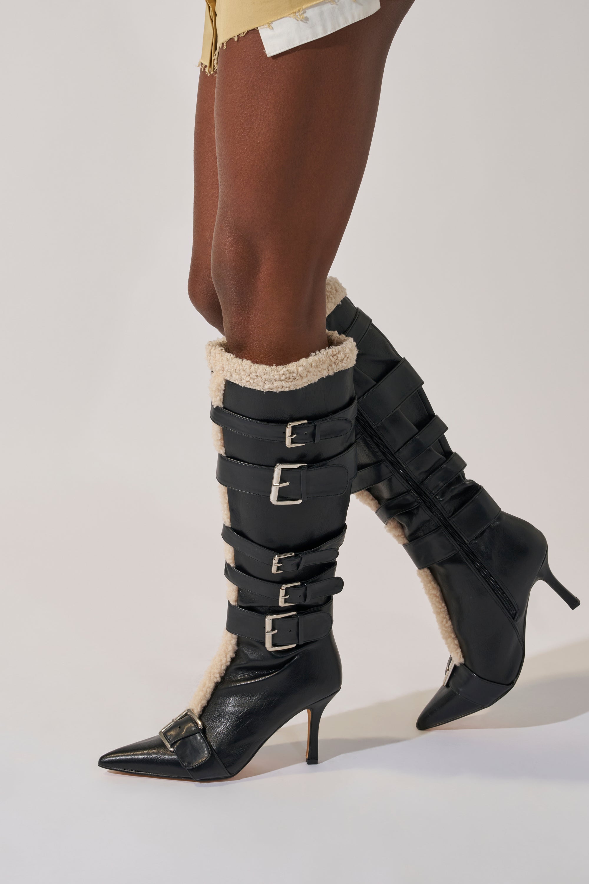 A person wears the AKIRA GOLD FIERO BLACK PU AND SHERPA BUCKLE knee-high boots with pointed toes, multiple silver buckles, and cream shearling trim, standing against a light background.