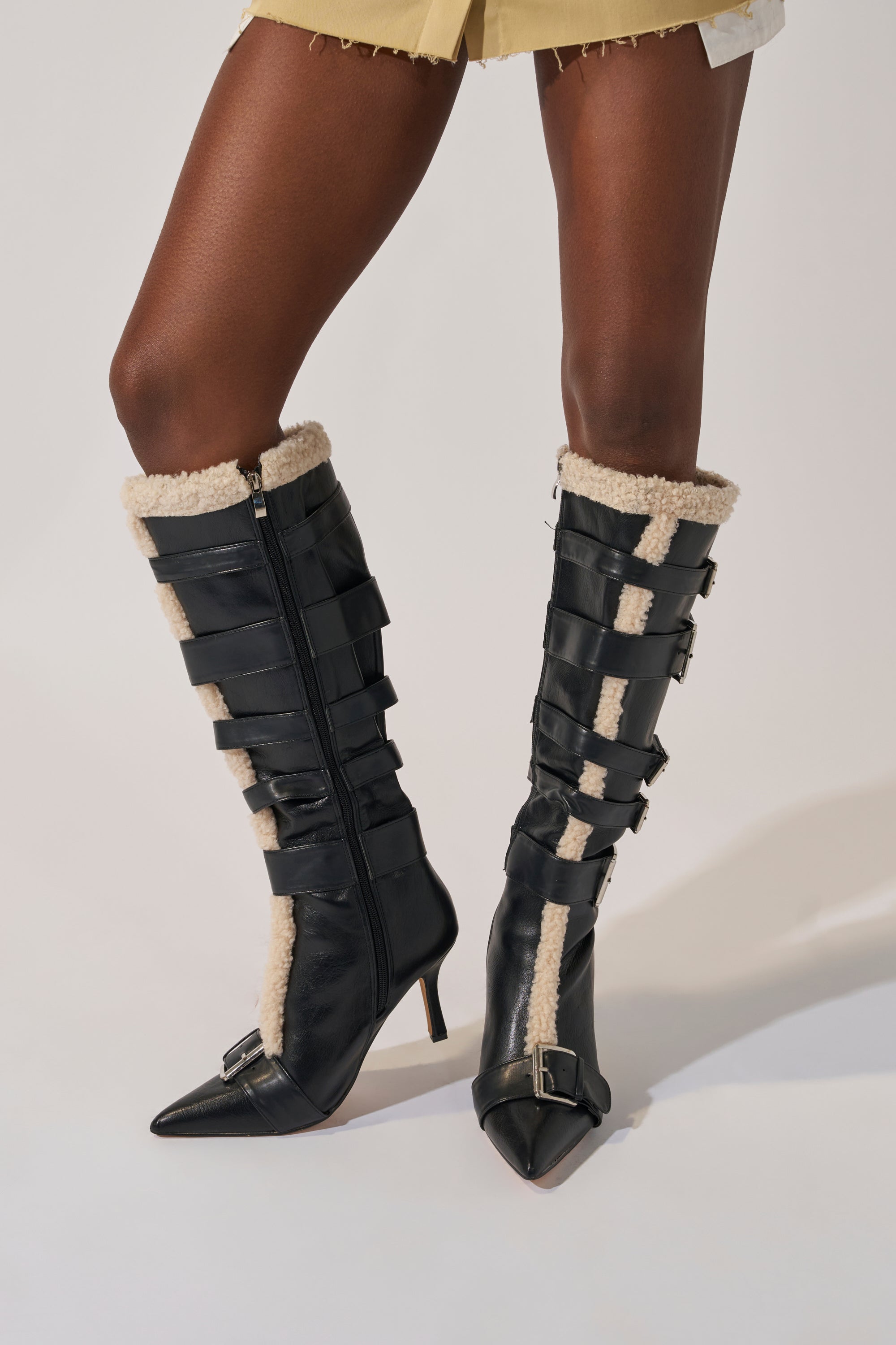 A person styles the AKIRA GOLD FIERO BLACK PU AND SHERPA BUCKLE boots—black faux leather with sherpa trim and multiple buckles, pointed toes, and stiletto heels—with a light-colored, frayed-hem skirt for a chic look.
