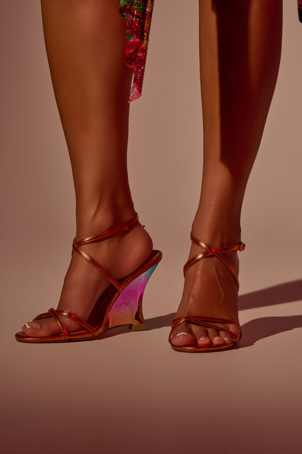 Someone stands on a neutral background wearing the AKIRA GOLD FINGA CHROME WEDGE STRAP SANDAL—metallic strappy wedges with a rainbow ombre heel—paired with a glimpse of a colorful outfit. 0