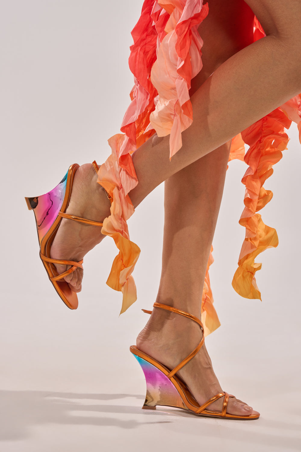 Wearing the AKIRA GOLD FINGA CHROME WEDGE STRAP SANDAL, a person in a ruffled pink and orange dress stands on tiptoe, showing off the sandals' strappy design and striking chrome wedge heels against a light background. 3