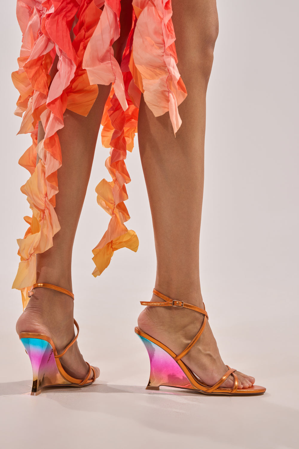 A person wears an orange ruffled skirt and AKIRA GOLD FINGA CHROME WEDGE STRAP SANDALS, standing on a light background. 8