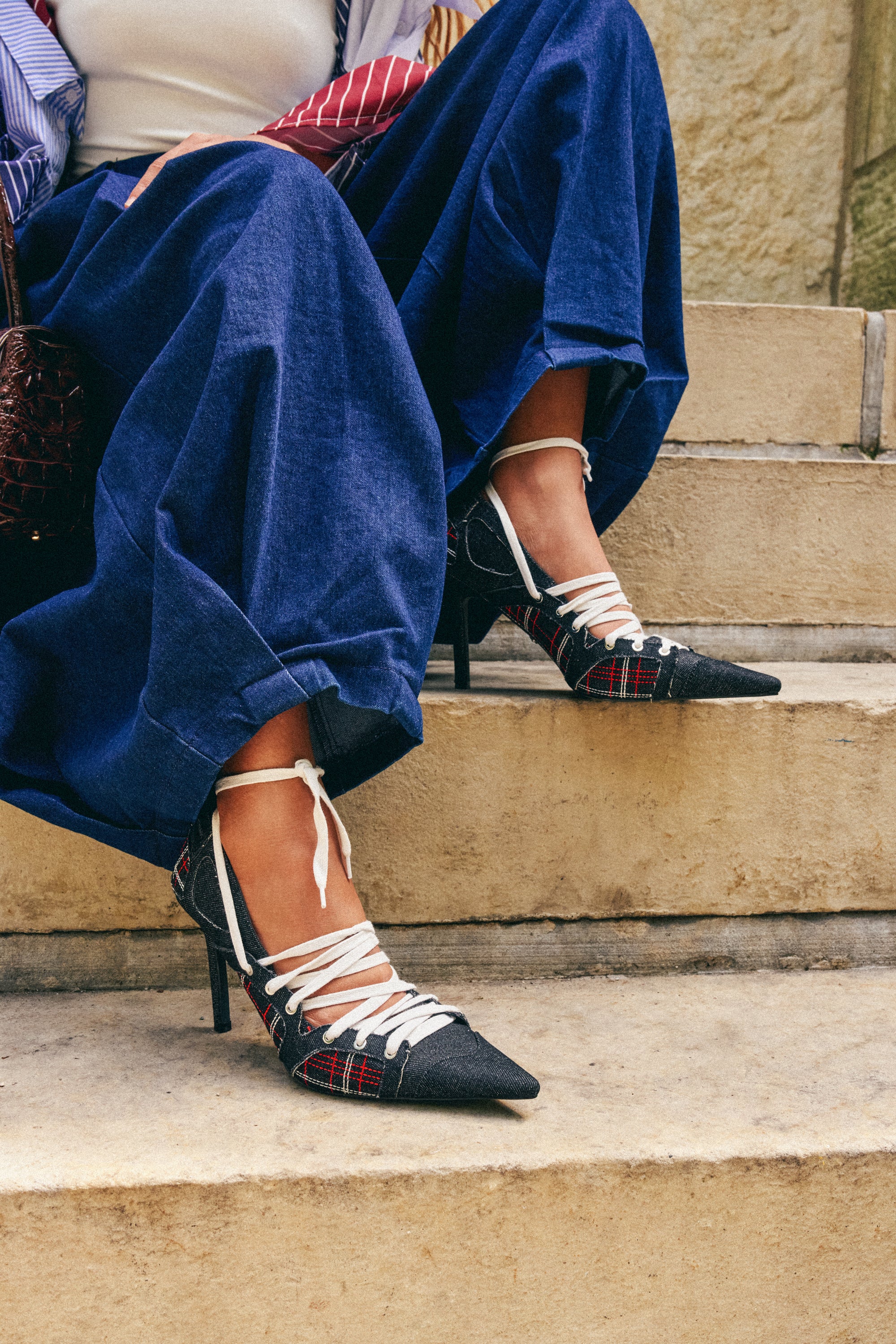 A person wearing a long blue skirt and the AZALEA WANG FIRE STRING PLAID SPORTY PUMP with pointed toes and white laces sits on stone stairs, legs crossed at the ankles.