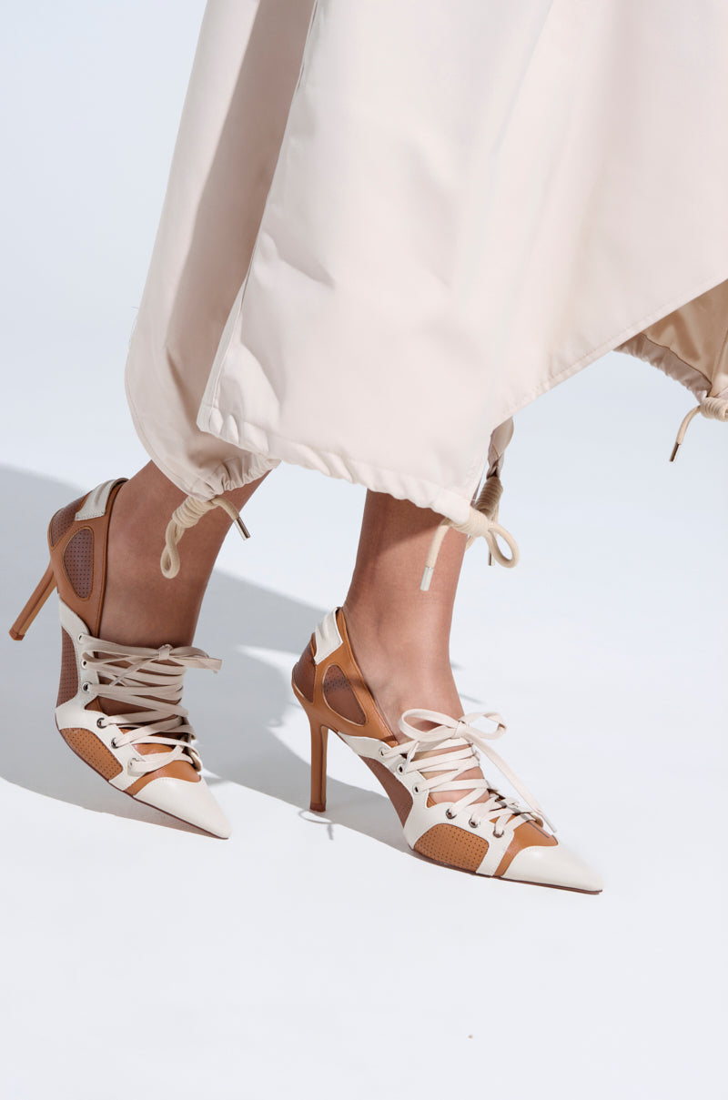 Someone is wearing the AZALEA WANG FIRST STRING TAN COLOR BLOCK SPORTY PUMP, featuring tan and white pointed high heels with sneaker-style laces. The lower part of a beige dress with adjustable drawstrings stands out against the plain light background.