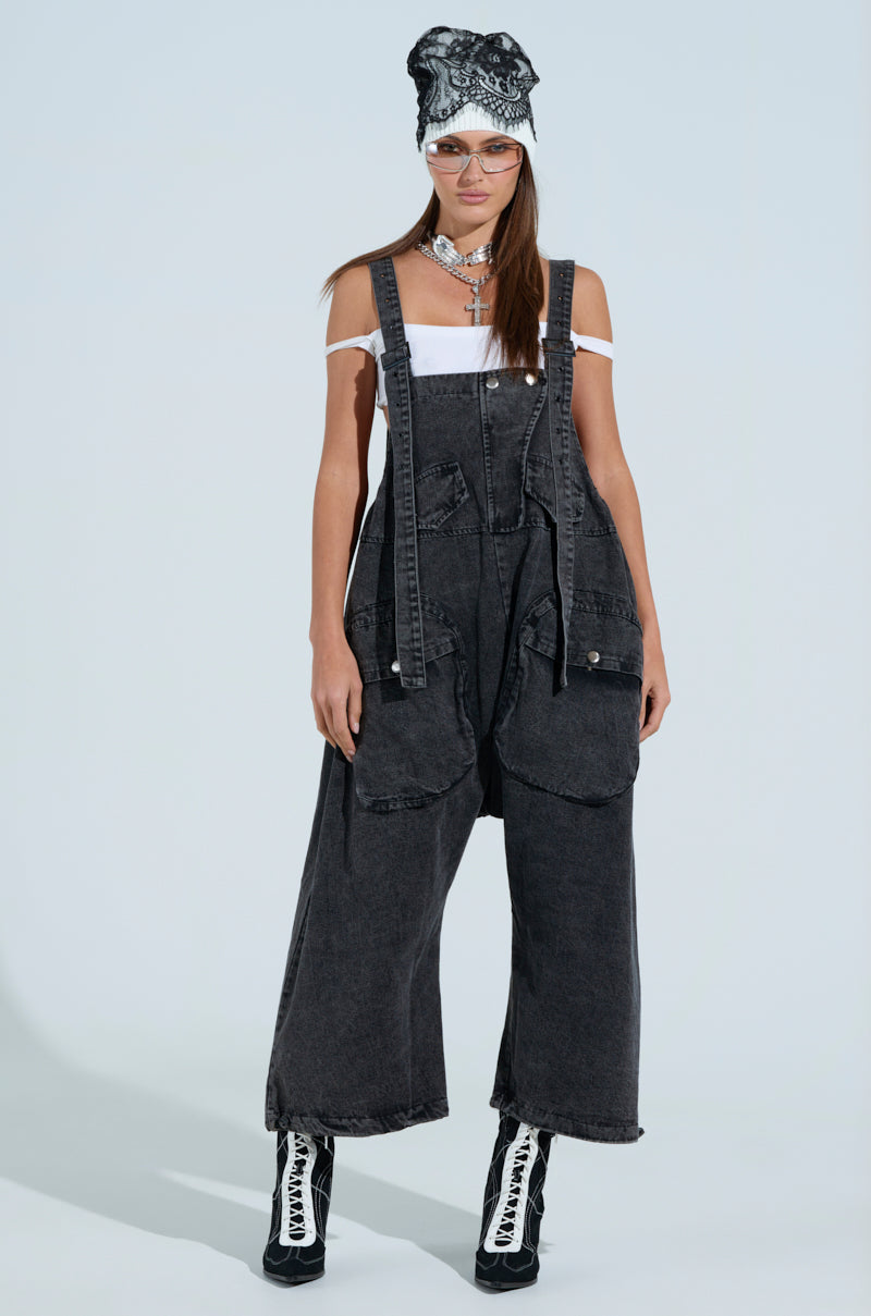 A woman flaunts a chic JUST LIKE THAT Baggy Fit Overall in grey with a white off-shoulder top and stylish black-and-white lace-up boots. She accessorizes with a patterned headwrap, glasses, and layered necklaces against a plain light-colored backdrop. The overall features an adjustable toggle hem for added flair.