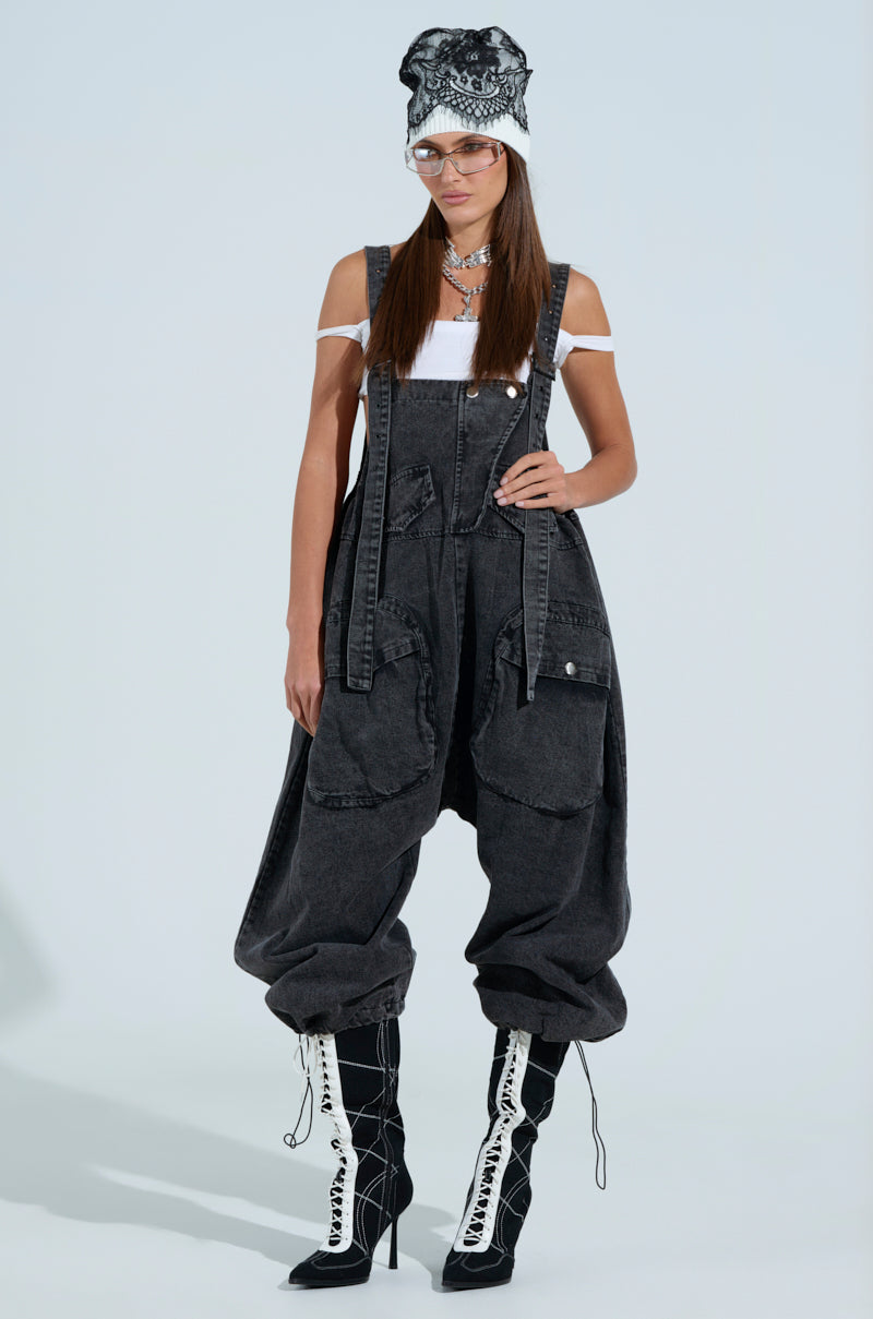 A person confidently wears a white off-the-shoulder top under "Just Like That Baggy Fit Overalls in Grey" with adjustable toggle hems, paired with a patterned beanie, glasses, and black-and-white lace-up high-heeled boots against a light background.