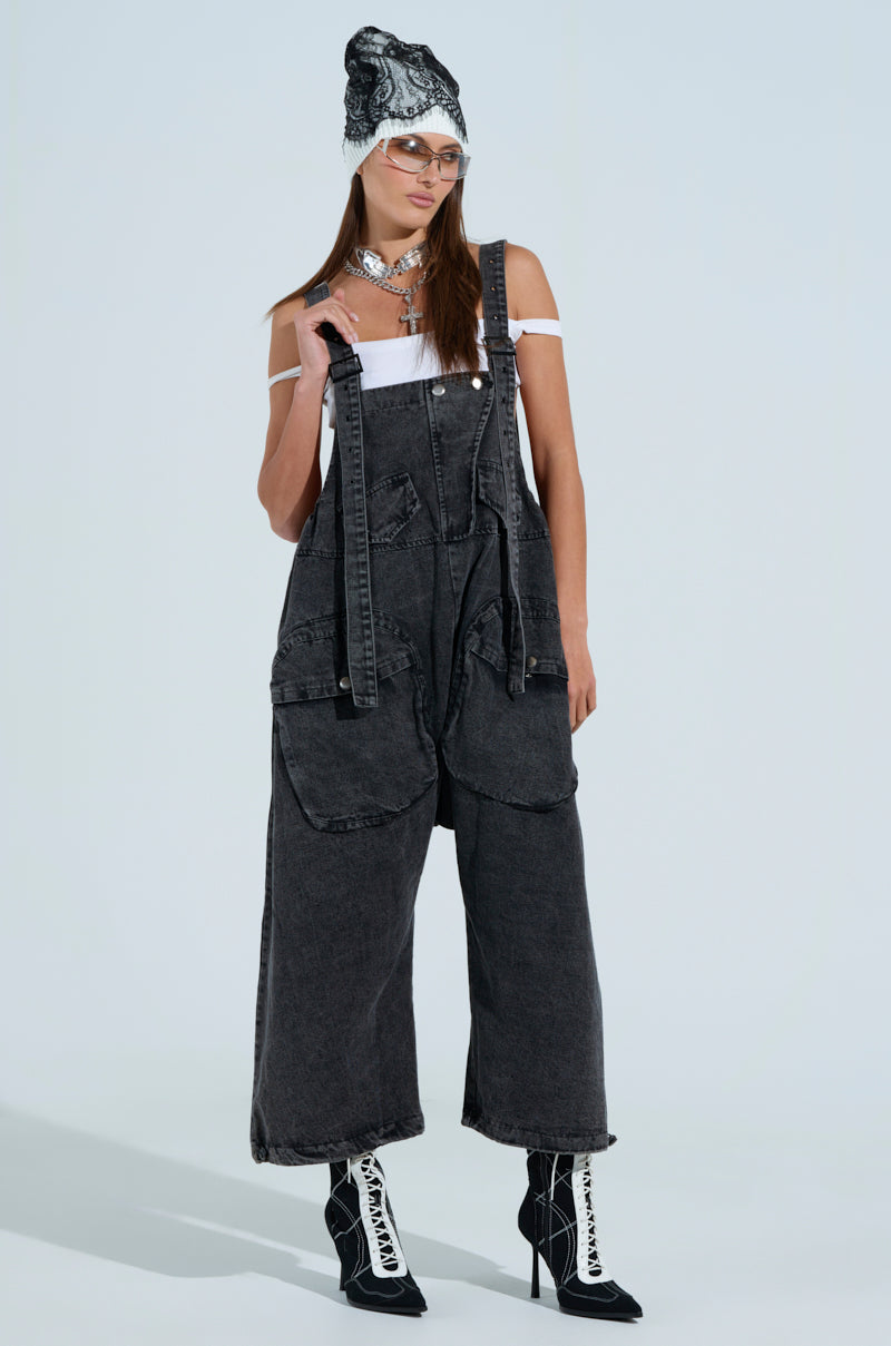 Wearing oversized grey baggy fit overalls, a white off-shoulder top, black pointed boots, a patterned hat, and clear glasses, layered necklaces adorn them as they stand against a plain background.