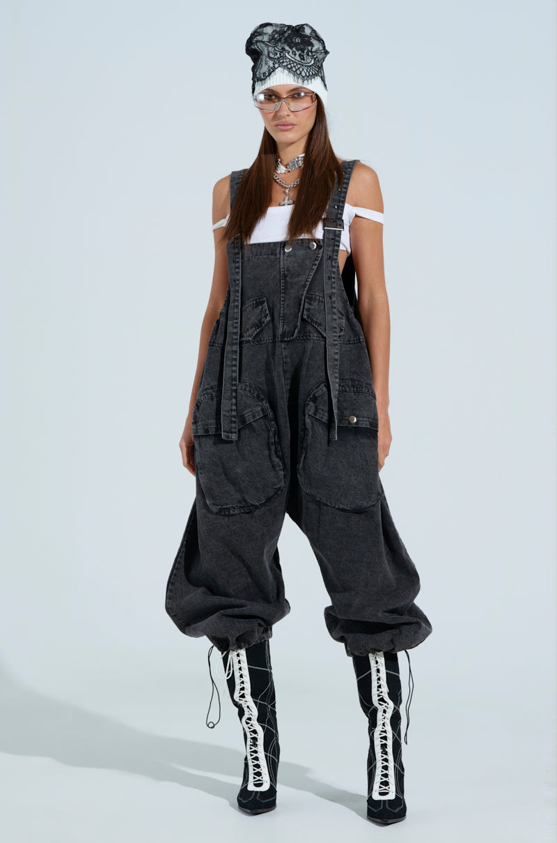 A person in the JUST LIKE THAT baggy fit overalls in grey with an adjustable toggle hem, a white off-shoulder top, and black lace-up boots. Accessories include a patterned beanie, clear glasses, layered necklaces against a light blue background.