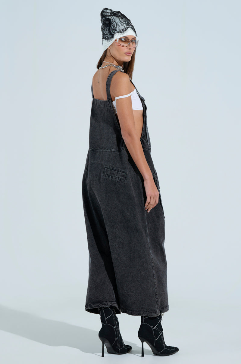 A person in grey baggy fit overalls, a white shirt, black patterned ankle boots, and a beanie poses against a light background with long, straight hair, gazing toward the camera.