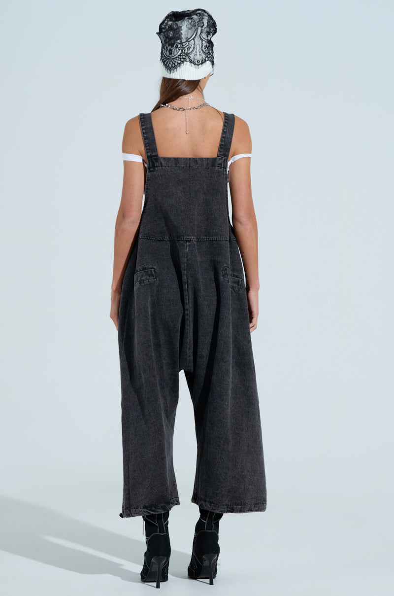 A person faces away, highlighting the "JUST LIKE THAT BAGGY FIT OVERALLS IN GREY" with wide legs and a white off-shoulder top. They have long hair, a black and white headscarf, paired with high-heeled shoes against a light blue background.