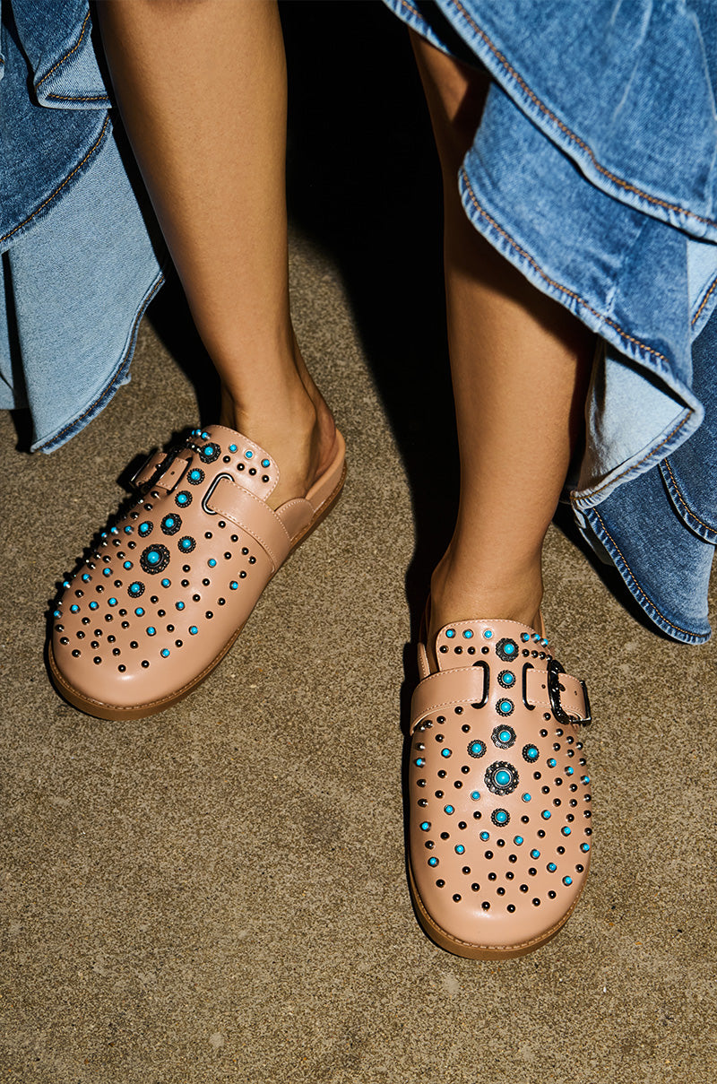Someone stands on a concrete floor wearing the AZALEA WANG FLAIR TAN EMBELLISHED CLOG with turquoise studs and buckles, paired with a blue ruffled denim skirt.