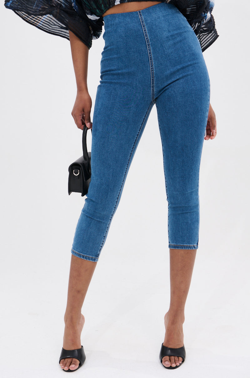 Wearing the FLEX FIT MEDIUM WASH CAPRI, a person in stretchy high-rise capris pairs them with black open-toe heels and holds a small black handbag, with part of a black patterned top visible.