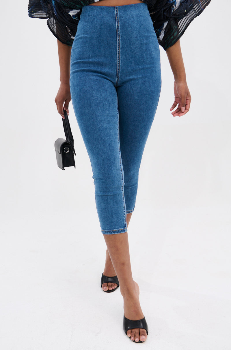 A person wearing the FLEX FIT MEDIUM WASH CAPRI in stretchy denim, styled with black open-toe heels and a small black handbag, walks forward against a plain white background.