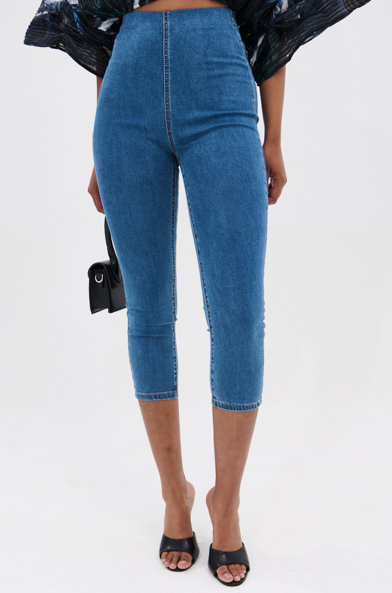 Wearing the FLEX FIT MEDIUM WASH CAPRI—high-rise, stretchy denim capri pants—a person pairs them with black mule sandals and a small black handbag, standing against a plain light background.