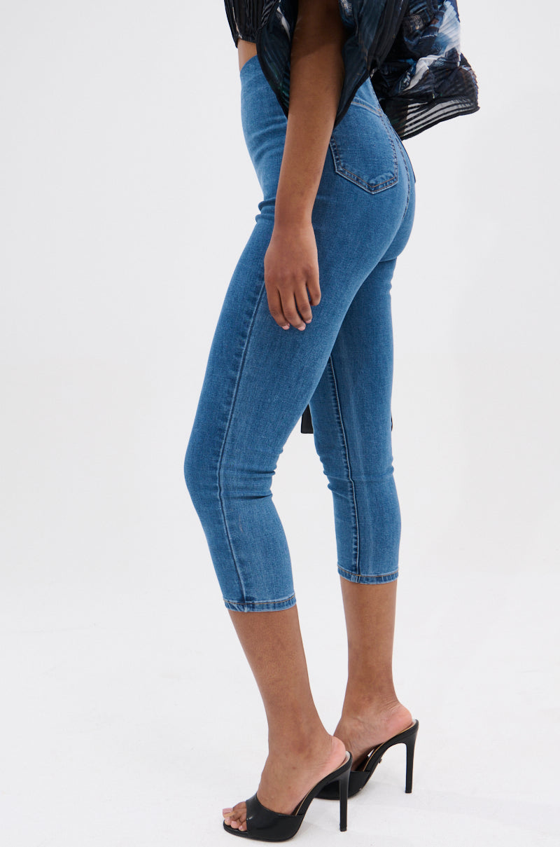 A person in the FLEX FIT MEDIUM WASH CAPRI—high-rise, blue cropped skinny jeans in stretchy denim—paired with a dark patterned top and black high-heeled mules poses against a plain light background, one leg slightly bent.