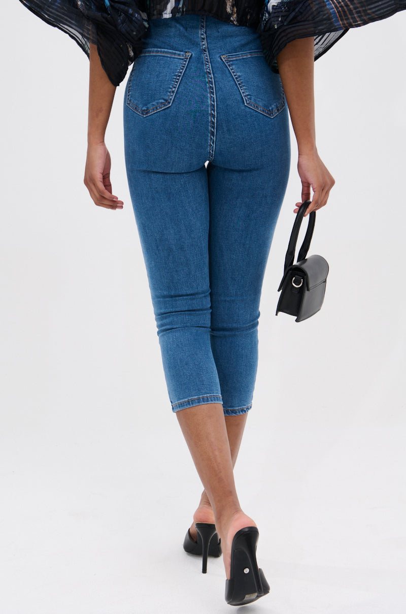 A person shown from the back against a light background wears FLEX FIT MEDIUM WASH CAPRI jeans, paired with black high heels and a small black handbag.
