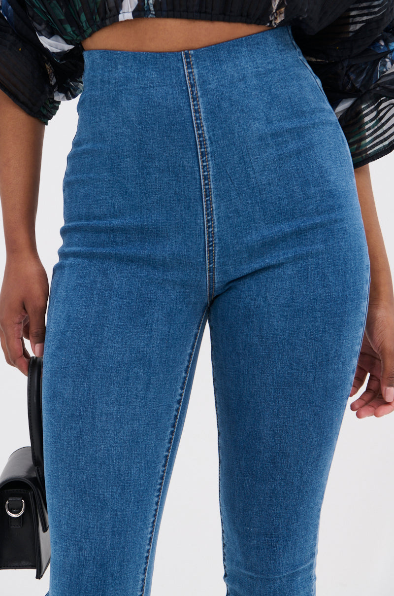 A close-up of a person wearing FLEX FIT MEDIUM WASH CAPRI jeans in stretchy denim, holding a small black handbag and showing a cropped patterned top above the jeans.