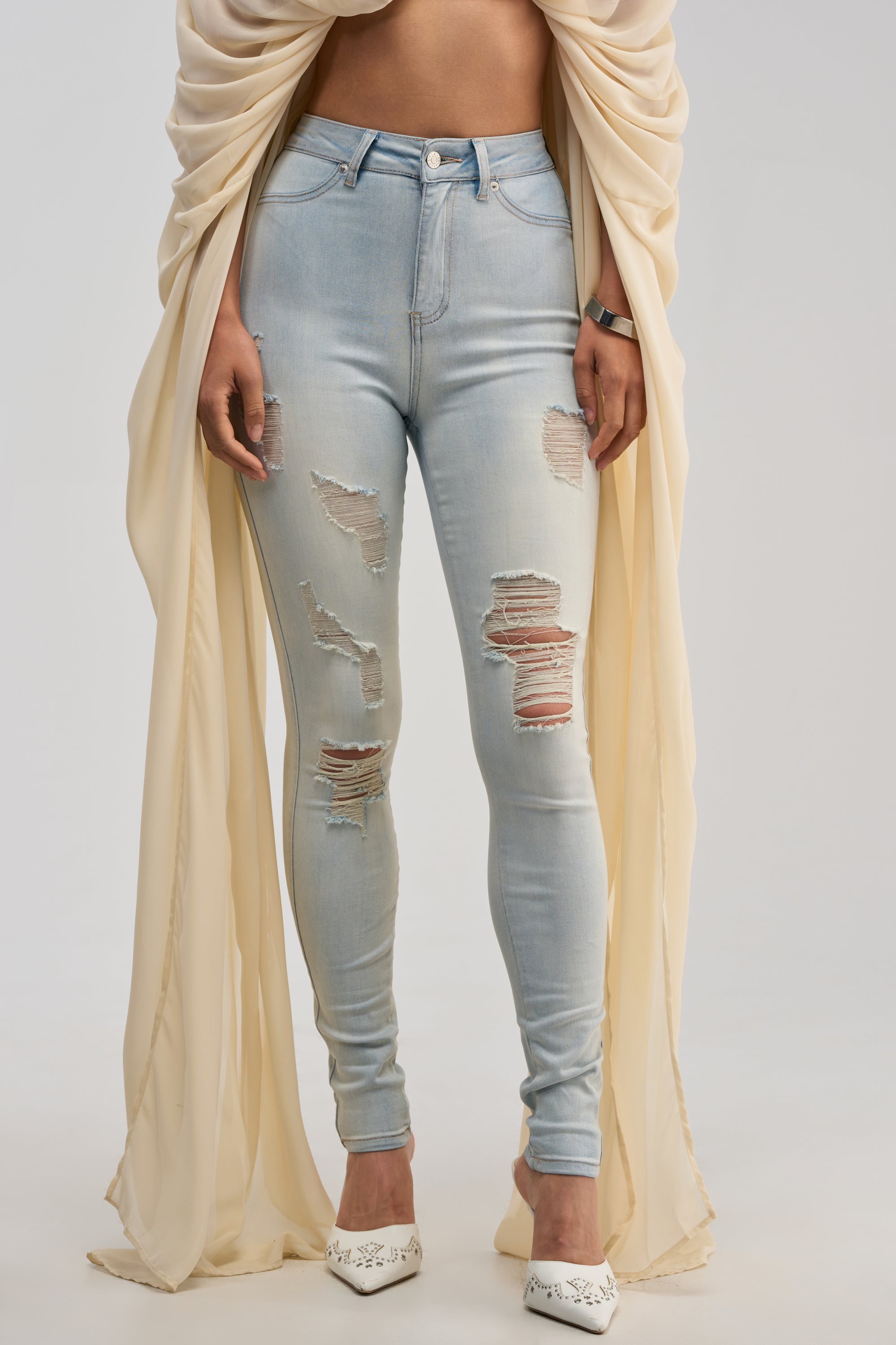 Wearing the FLEX-FIT EXTREME STRETCH HIGH WAISTED DISTRESSED SKINNY JEANS in a light wash, paired with a cream draped top, a black watch, and white pointed shoes with small designs, this individual stands against a simple light background.