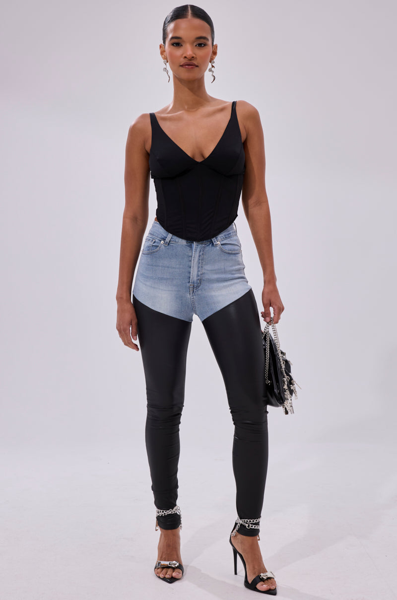 A woman stands confidently in a studio wearing FLEX FIT EXTREME STRETCH HIGH WAIST SKINNY JEANS WITH FAUX LEATHER, a black sleeveless top, black leggings, high-heeled sandals, hoop earrings, and holding a chain-detail handbag.