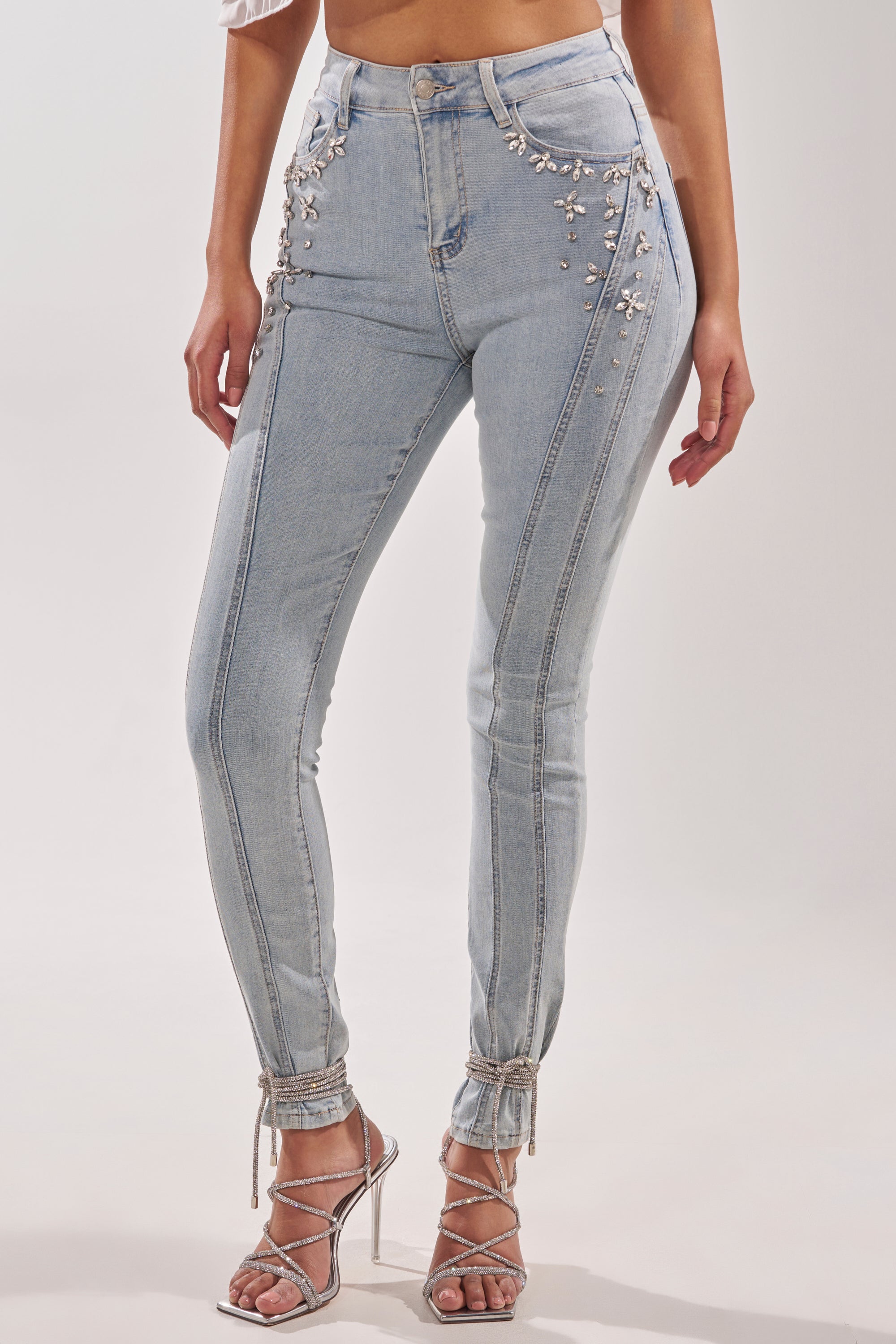 A woman wearing FLEX FIT SKINNY DENIM PANTS WITH RHINESTONES, light-wash and high-rise, paired with a cropped top and strappy silver heels. Only her torso and legs are visible.