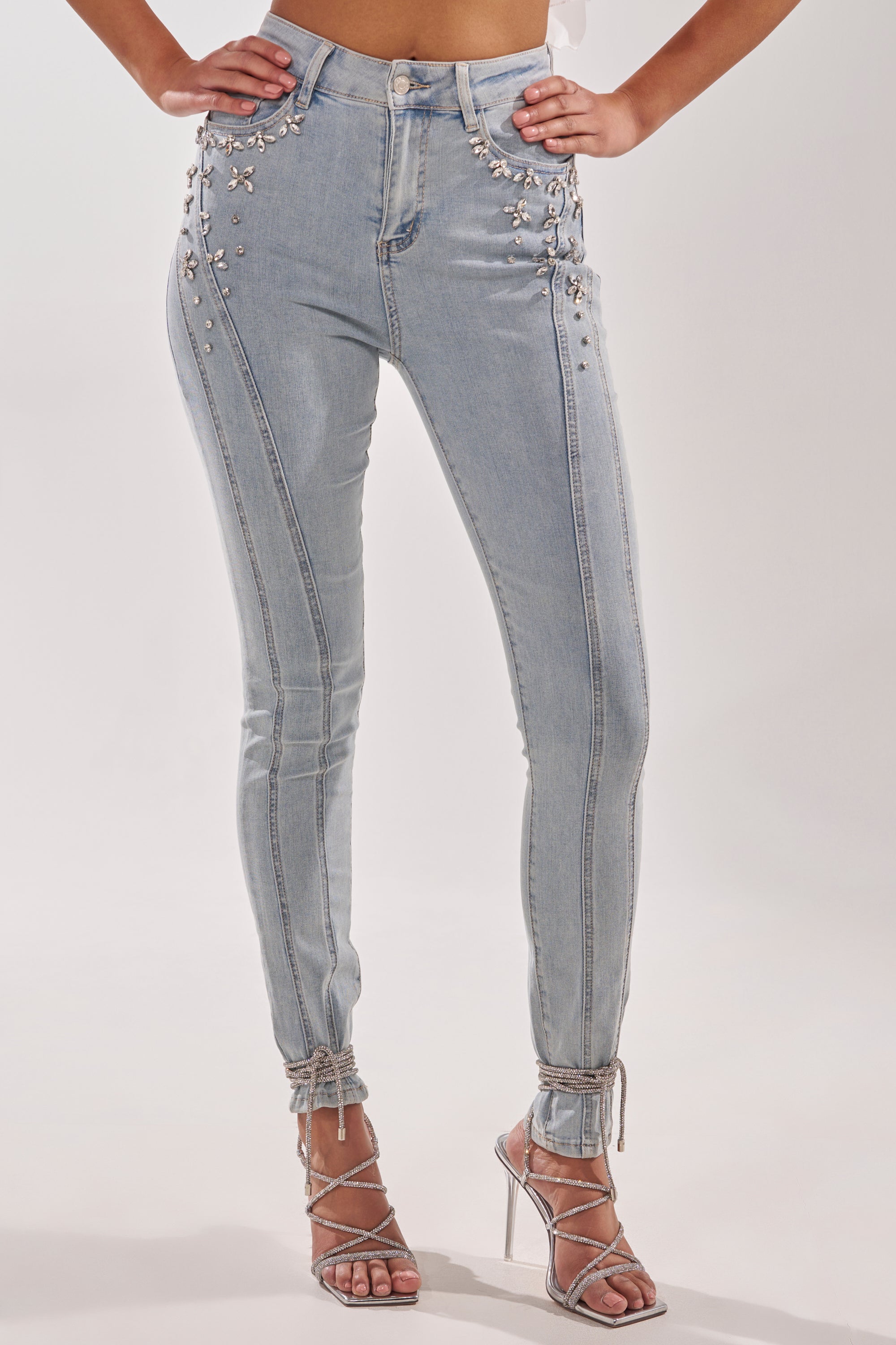 A woman models FLEX FIT SKINNY DENIM PANTS WITH RHINESTONES, featuring high-rise styling and silver rhinestone embellishments on the front pockets and side seams. She pairs them with strappy high heels against a plain white background.