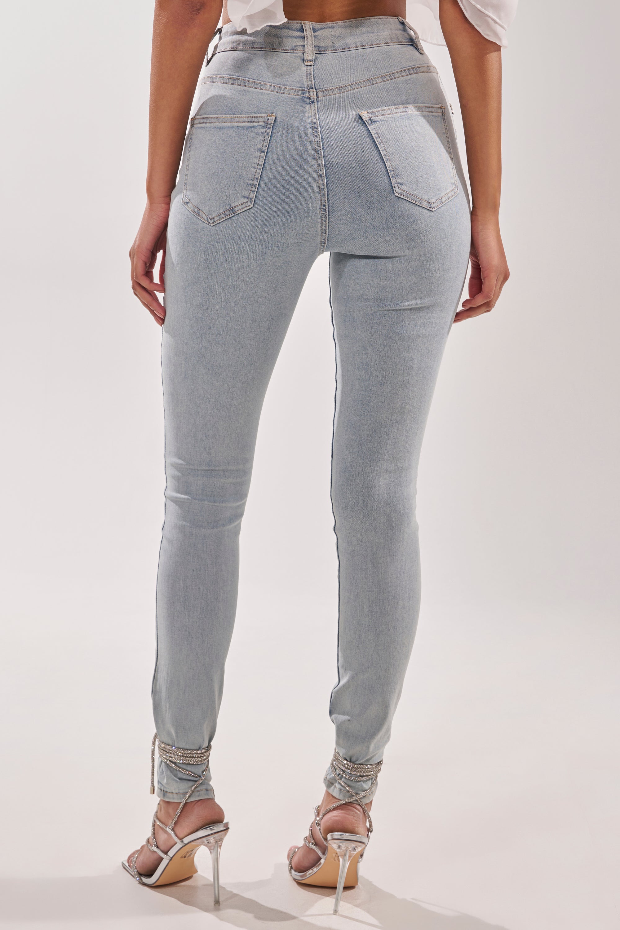 A person is seen from the back wearing FLEX FIT SKINNY DENIM PANTS WITH RHINESTONES and strappy high-heeled sandals, standing against a plain light background.