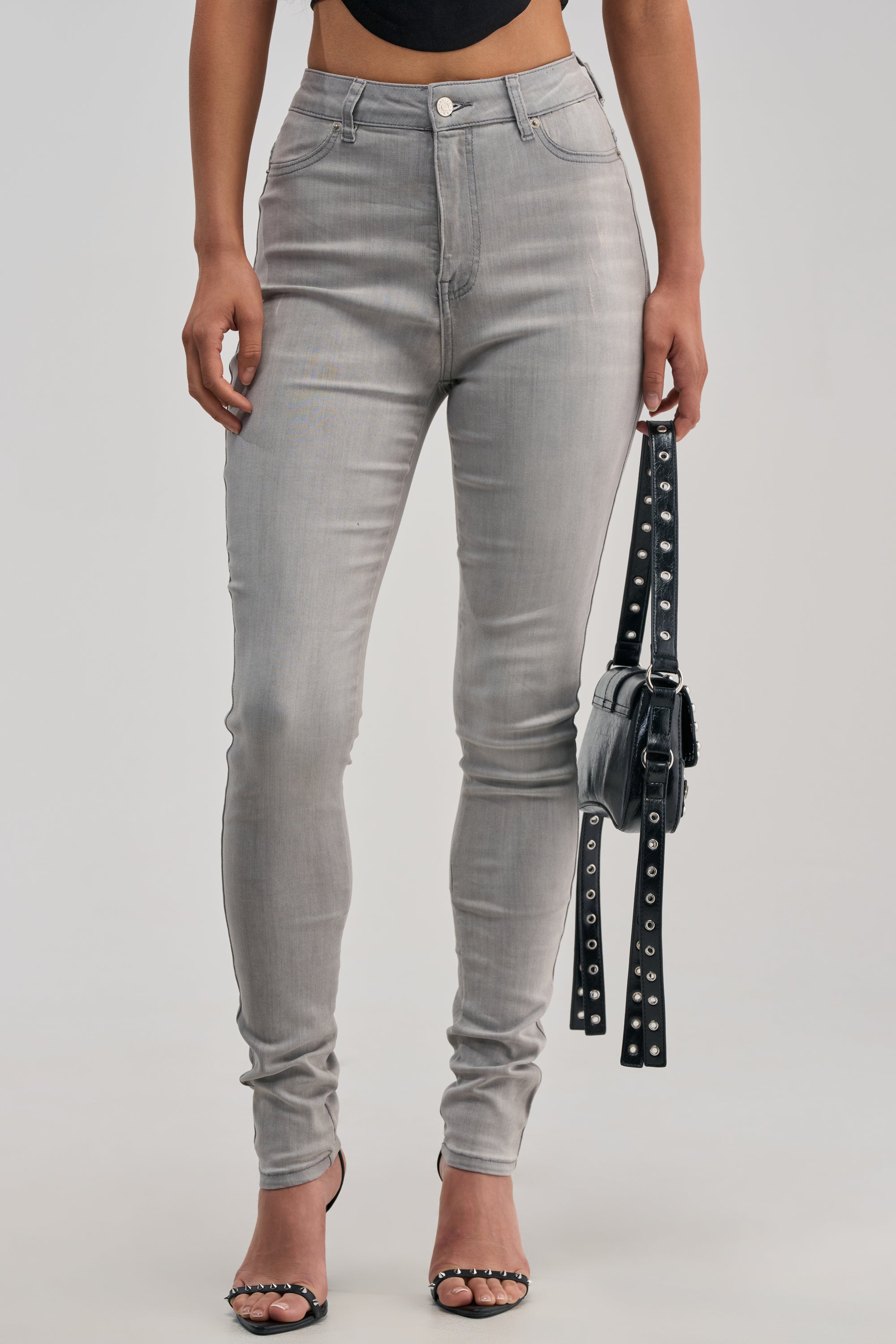 Wearing the FLEX-FIT High Waisted Super Stretchy Skinny Jeans in Grey, a person pairs their look with a black crop top, black high-heeled sandals, and carries a small black bag featuring silver studs and buckles.
