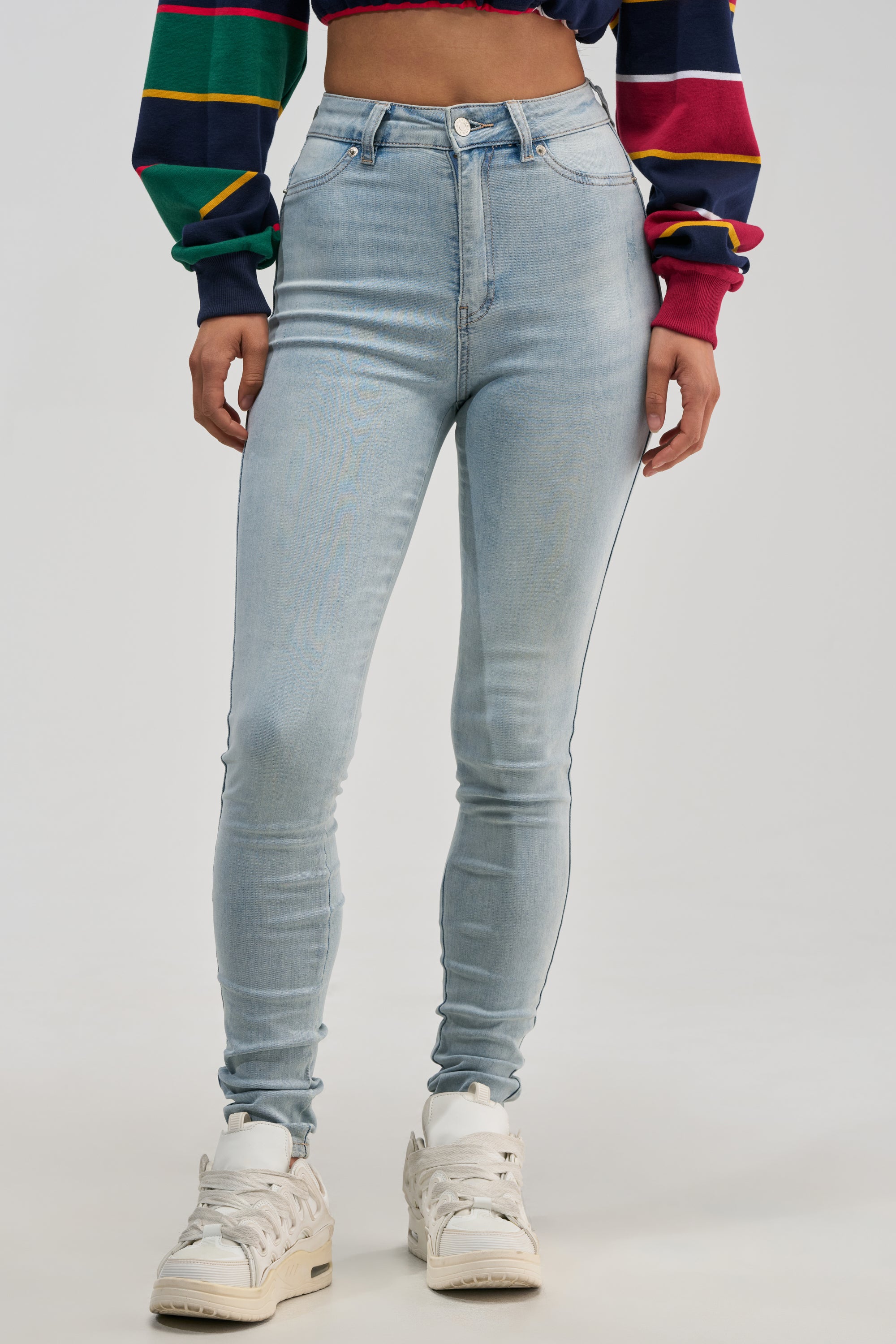 A person wearing FLEX-FIT HIGH WAISTED SUPER STRETCHY SKINNY JEANS IN LIGHT BLUE, paired with a colorful striped long-sleeve crop top and white sneakers, stands against a plain light background.