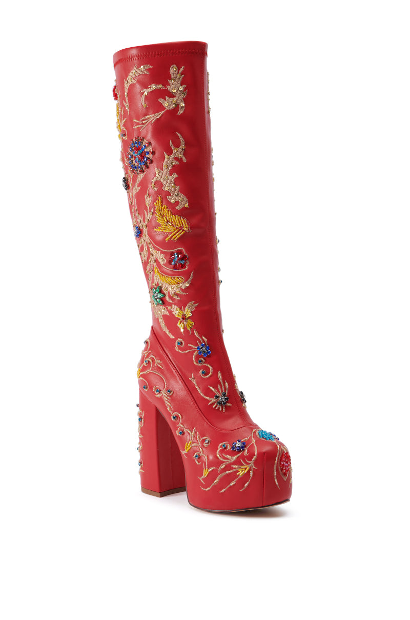 The AZALEA WANG FLIPPER RED PU WITH EMBELLISHMENTS CHUNKY BOOT is a red knee-high boot with a high platform heel, adorned with intricate, colorful floral embroidery. The design features a variety of embroidered flowers, leaves, and decorative patterns in vibrant hues on faux leather, creating a visually striking and embellished look.