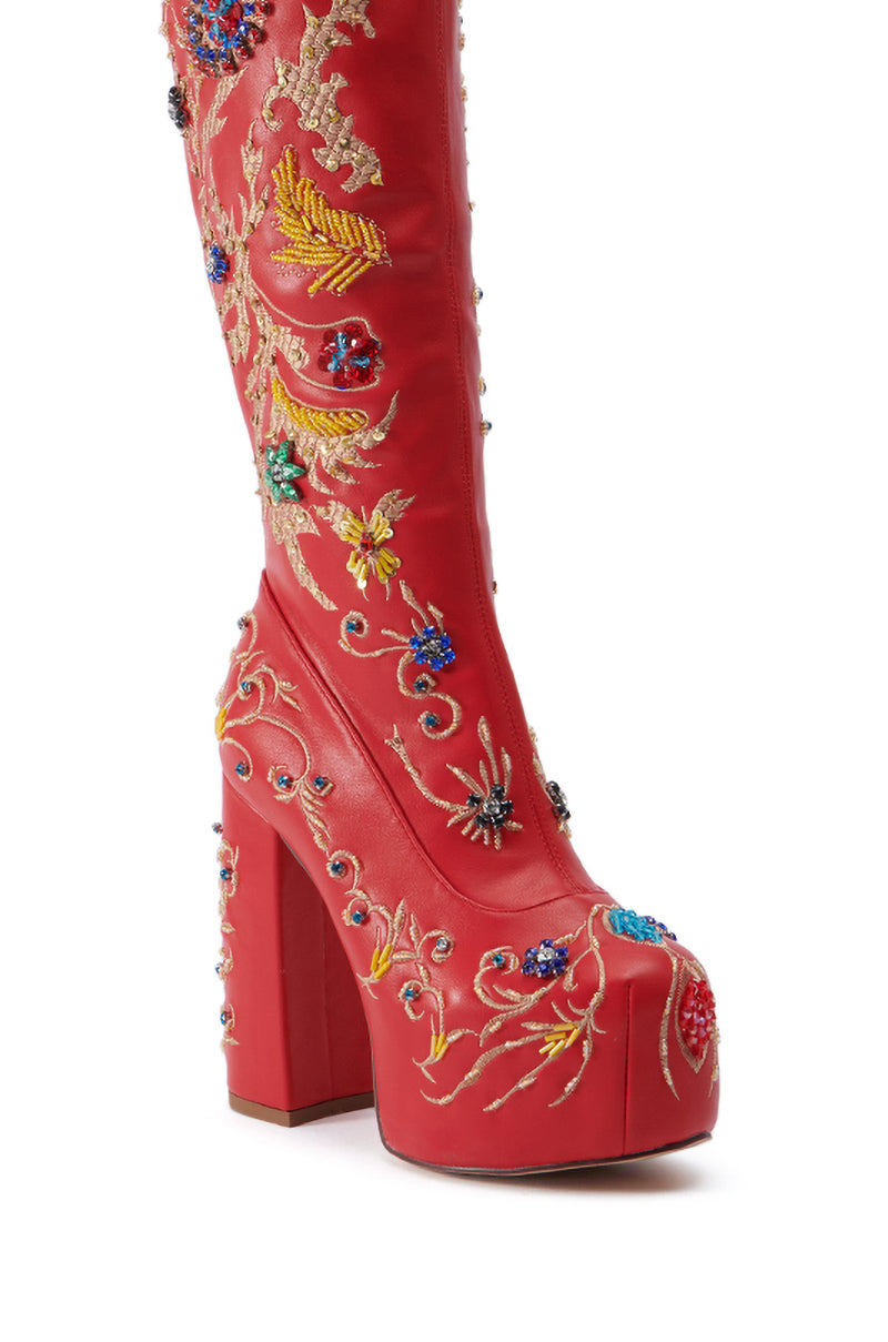 The AZALEA WANG FLIPPER RED PU WITH EMBELLISHMENTS CHUNKY BOOT is a knee-high platform boot in red faux leather, beautifully adorned with colorful embroidered flowers, insects, and ornamental patterns. It features a high chunky heel and a rounded toe.