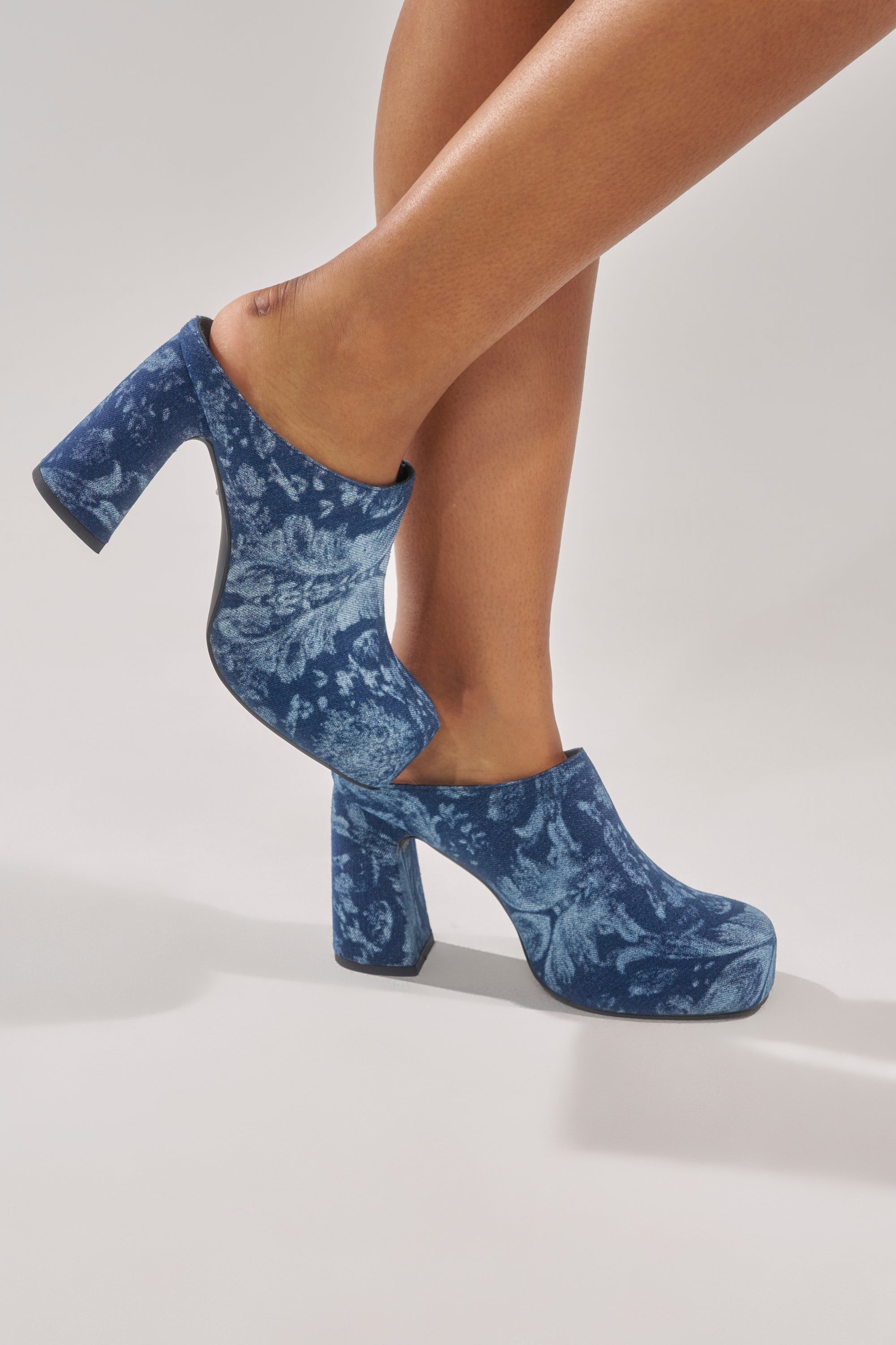 A person models the AZALEA WANG FLOREA DENIM PRINT MULE—blue, high-heeled slip-on mules with a floral pattern—one foot slightly raised to highlight the shoes against a light background. 1