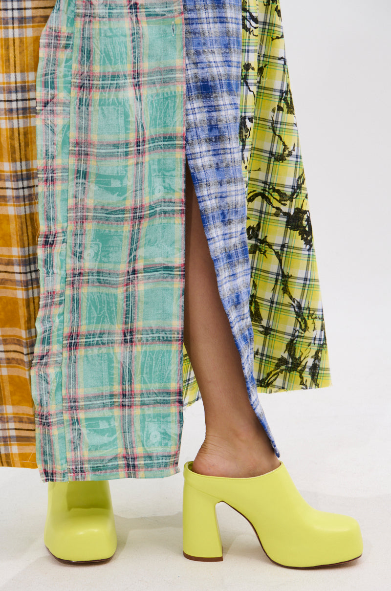 A person stands on a white background in a long, multicolored patchwork skirt with plaid and tie-dye patterns, revealing one leg through a slit. They wear the AZALEA WANG FLOREA LIME CLOG STYLE PUMP on their feet.