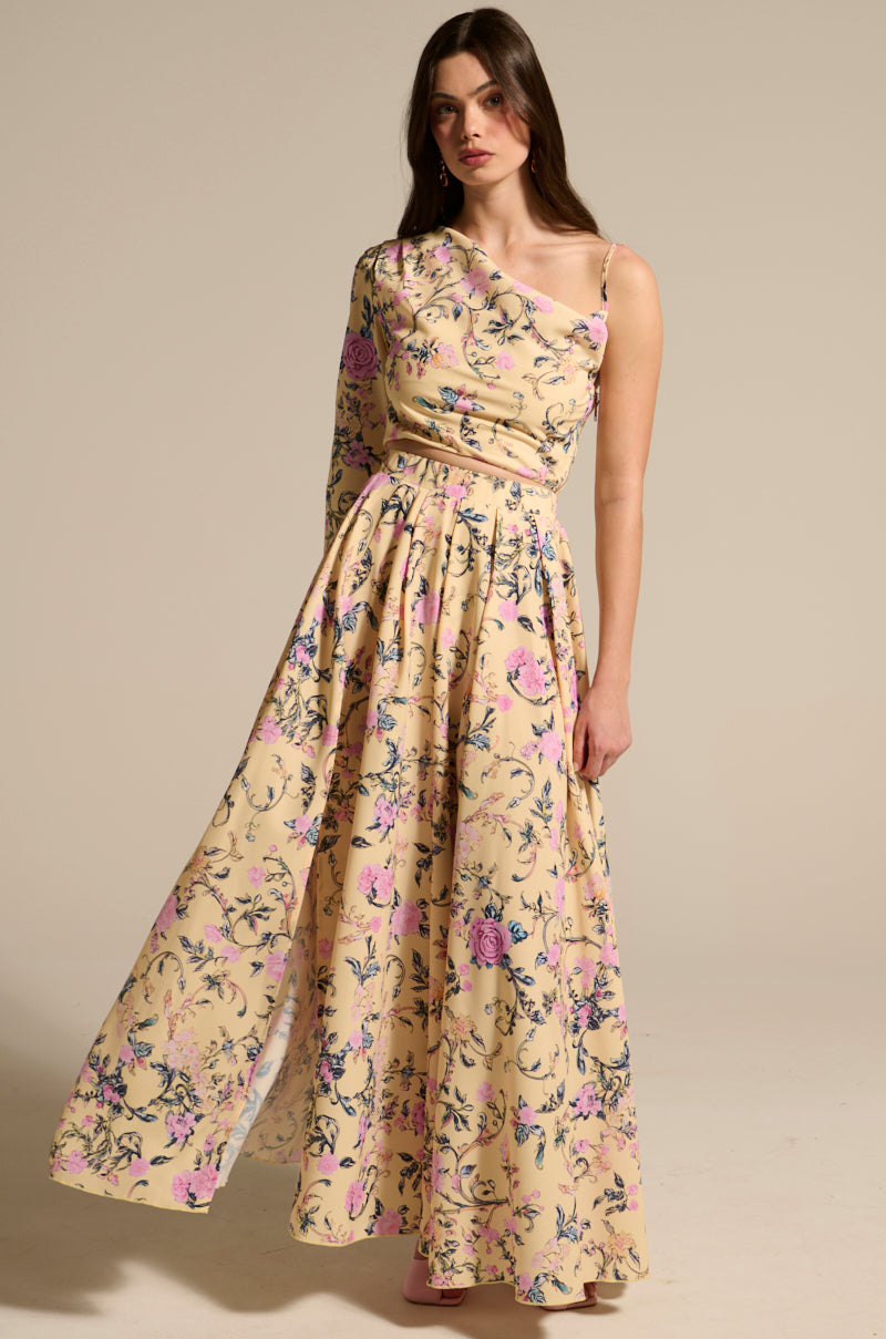A woman stands against a plain backdrop wearing the JOAN MAXI SKIRT—a high-waisted, floral print maxi skirt with a beige background, one long sleeve, and an off-the-shoulder design.