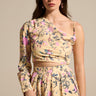 A woman models the JOAN BLOUSE, a cream floral one-shoulder blouse with pink and blue flowers, long sleeve, cropped hem, and a matching high-waisted skirt featuring a thigh-high slit.