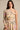 A woman with long brown hair models the yellow floral JOAN BLOUSE—one-shoulder with one long sleeve and one thin strap—paired with a high-waisted skirt, standing against a plain beige background.