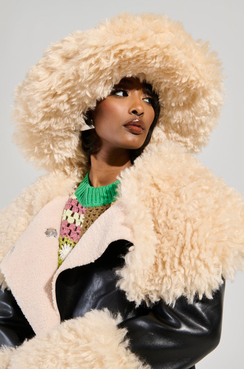 A person wearing the chic ULTIMATE FLUFF BUCKET HAT and a matching ivory fur coat with a black leather base layered over a vibrant crocheted top. The coat features a large collar, and they gaze to the side against a plain backdrop, their ensemble radiating fuzziness and sophistication.