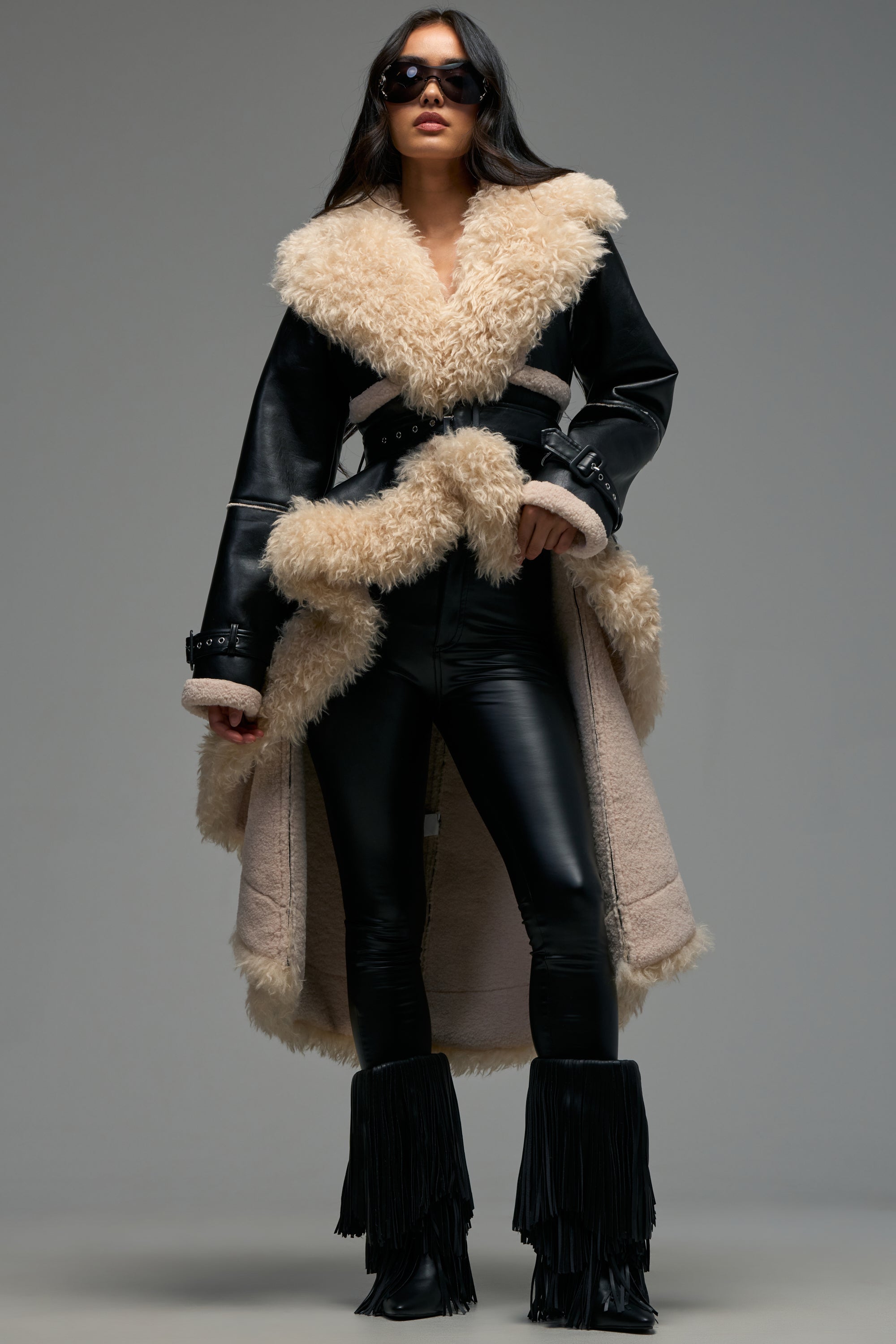 Wearing the TAKE YOU THERE FUR TRIM HIGH LOW TRENCH—a long black leather coat with a wide tan shearling collar and lining—this woman pairs it with black leather pants, black fringe boots, and sunglasses as she stands confidently against a plain background.