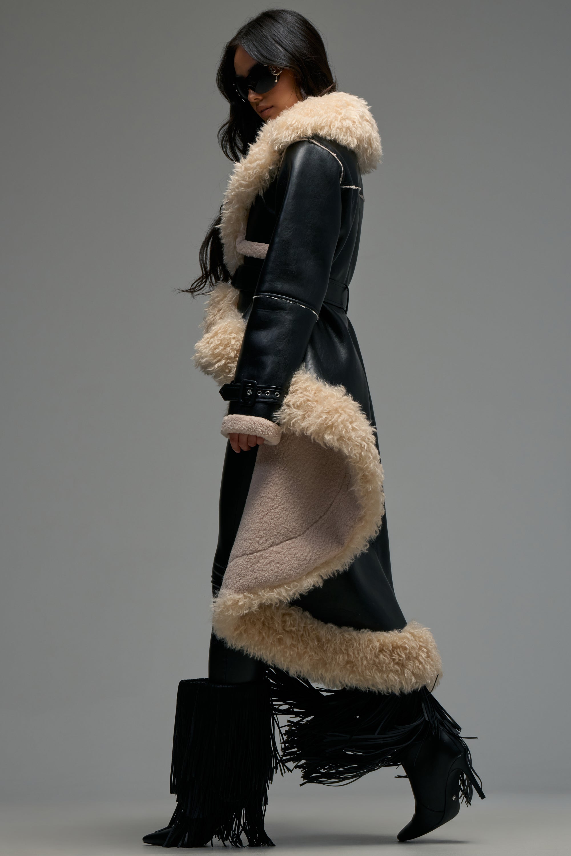 Wearing the TAKE YOU THERE FUR TRIM HIGH LOW TRENCH—a long black leather coat with tan shearling trim—a woman pairs it with black fringe boots and sunglasses as she walks confidently against a plain gray background.