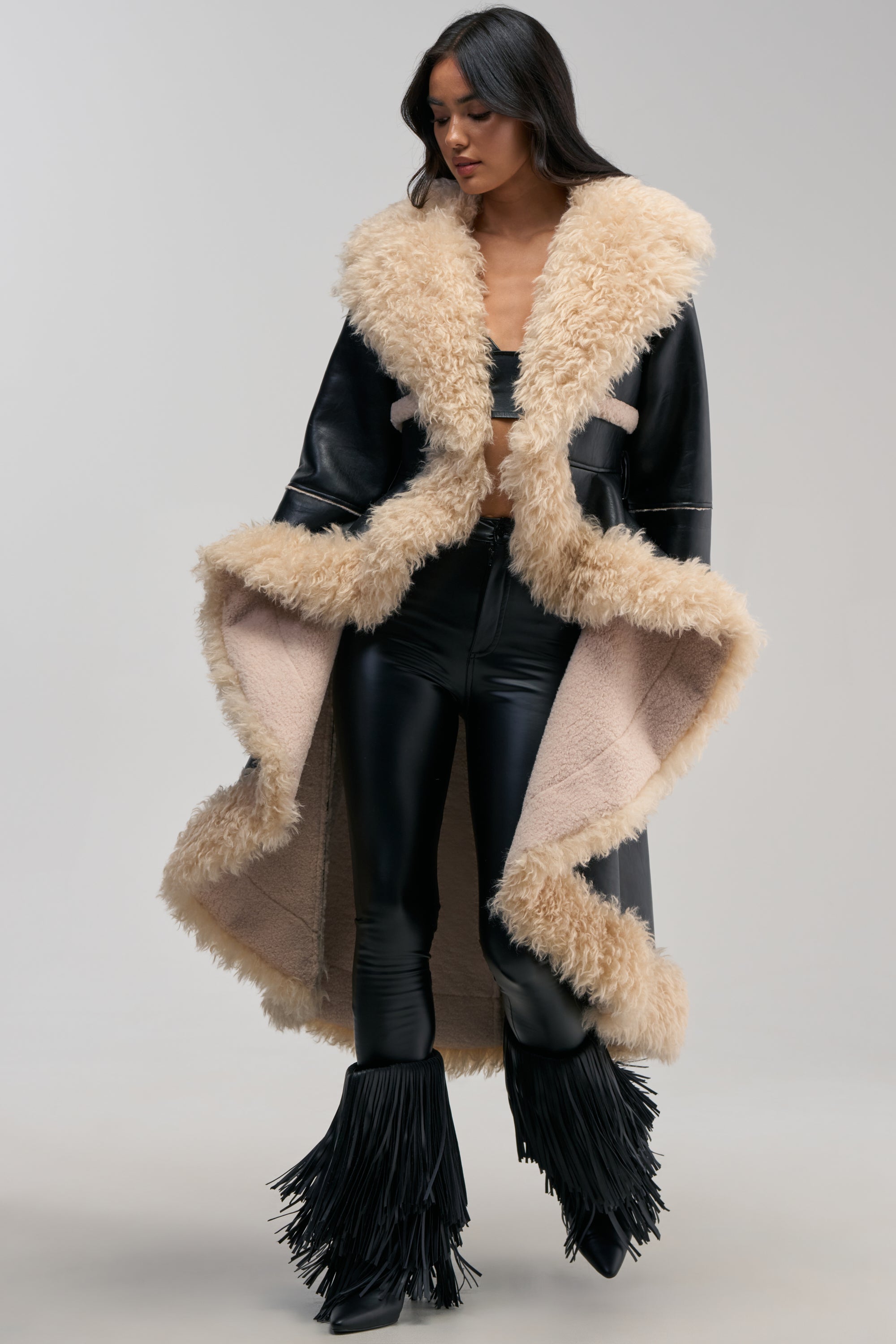 Wearing the TAKE YOU THERE FUR TRIM HIGH LOW TRENCH—a long black coat with beige shearling trim—a woman in black leather pants, a black bralette, and black fringe boots stands against a plain background.
