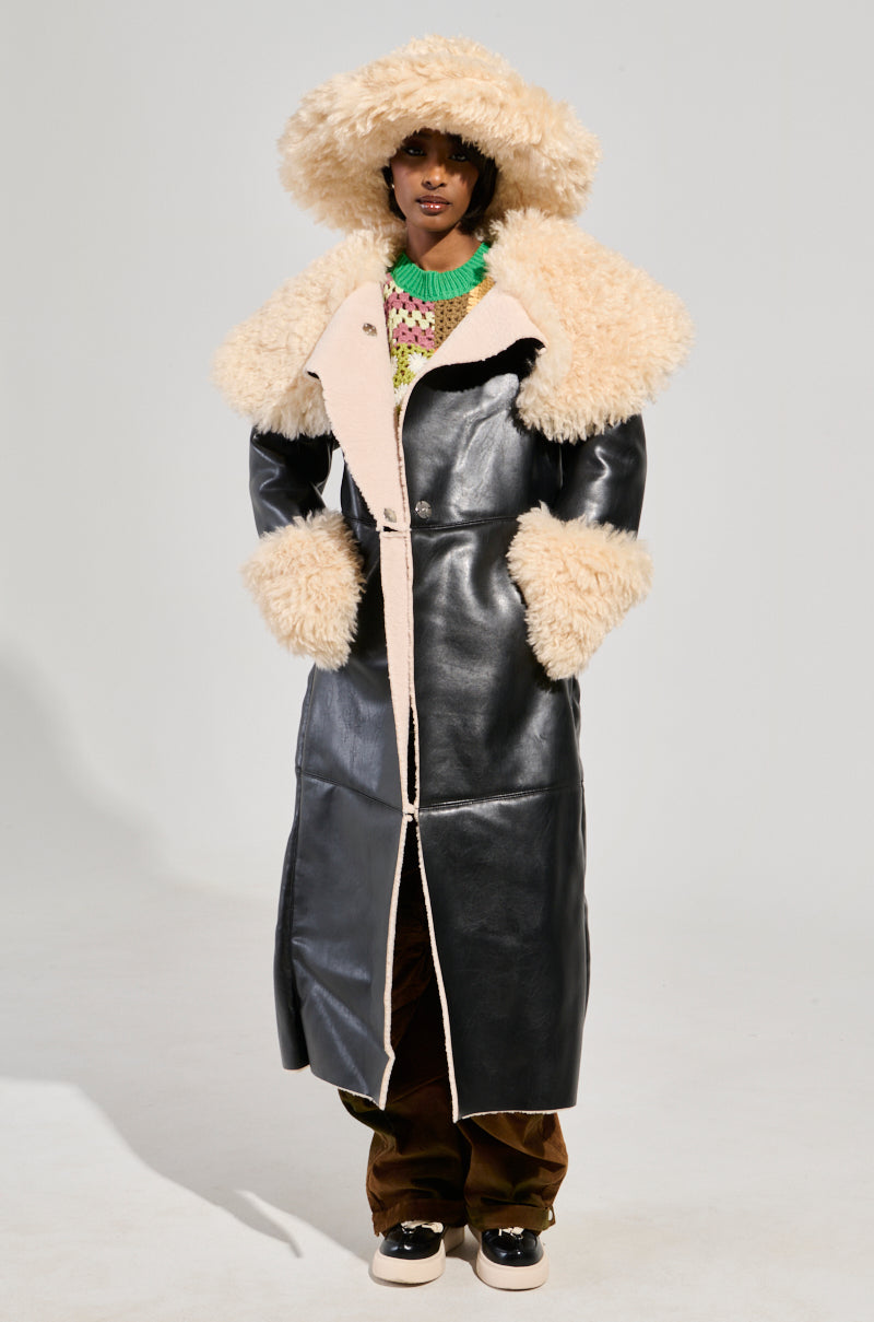 A person is wearing the SHE GOT IT FLUFFY PU TRENCH WITH BONDED SHERPA, a long black faux leather trench coat featuring a fluffy Sherpa collar and cuffs, paired with a wide-brimmed hat. They have on brown pants and sneakers to complete the ensemble, with a colorful knitted top visible underneath. The background remains plain.